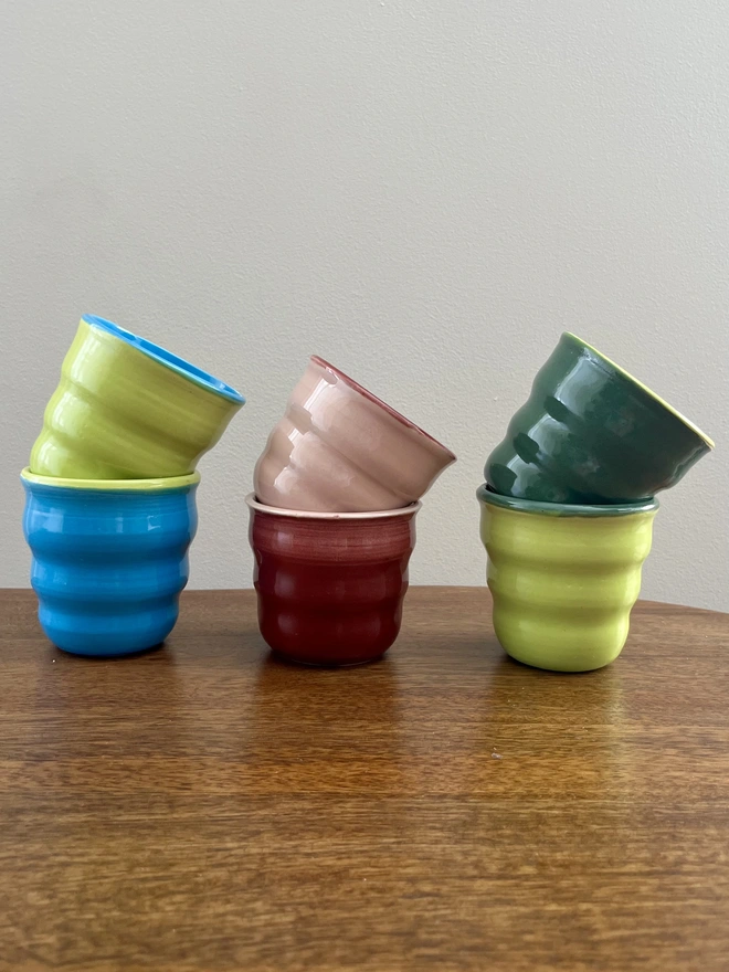 a row of 6 mini ceramic tumblers with wavy sides on a wooden table, they are stacked in matching pairs. each pair is glazed in the reverse colours.