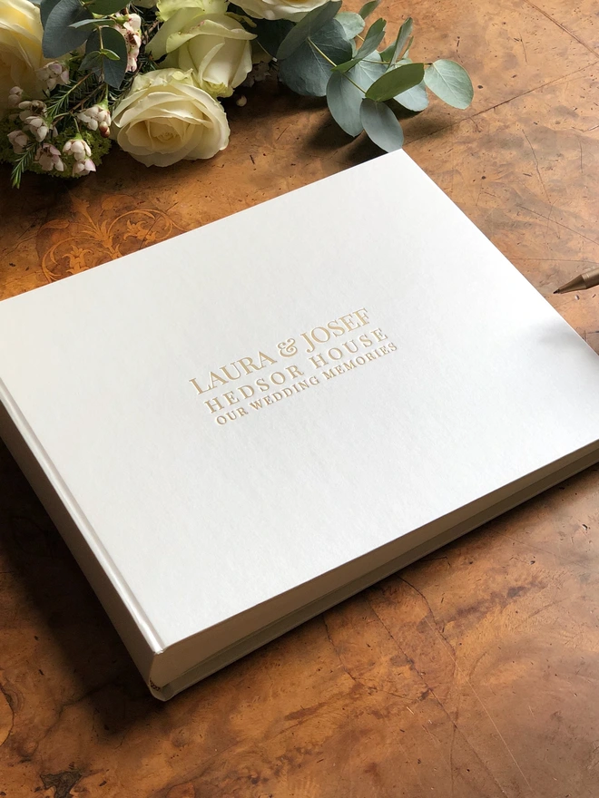 an ivory wedding guest book is lying on a beautiful walnut table ready to be signed. There are wedding flowers in the background.