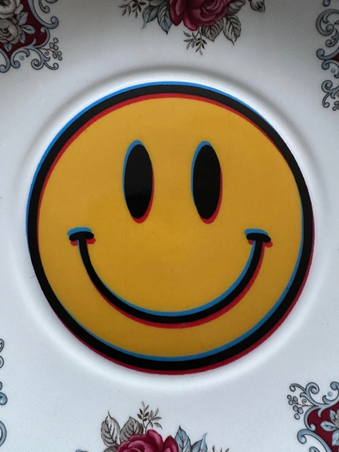 A round plate with a border of roses and a yellow smiley face in the center