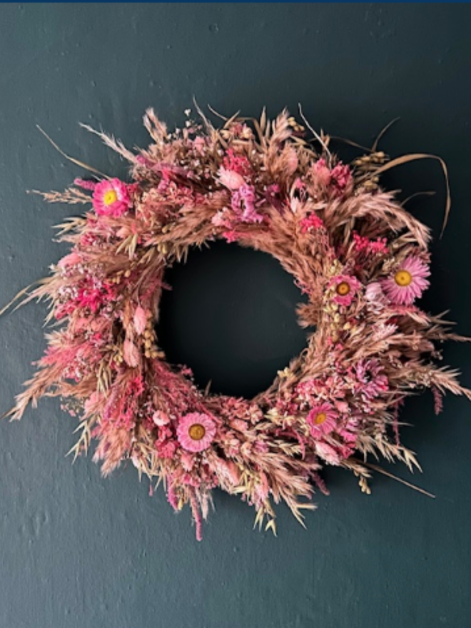 Pink Dried Floral Wreath. a circular wreath made of pink dried flowers hung up on a forest green wall.