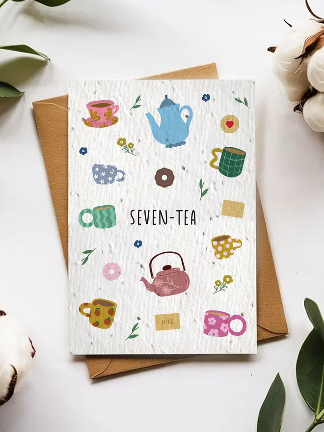 A greeting card titled “SEVEN-TEA” featuring colorful illustrations of teapots, mugs, flowers, and tea bags, placed on top of a brown envelope surrounded by cotton flowers and green leaves on a white background.