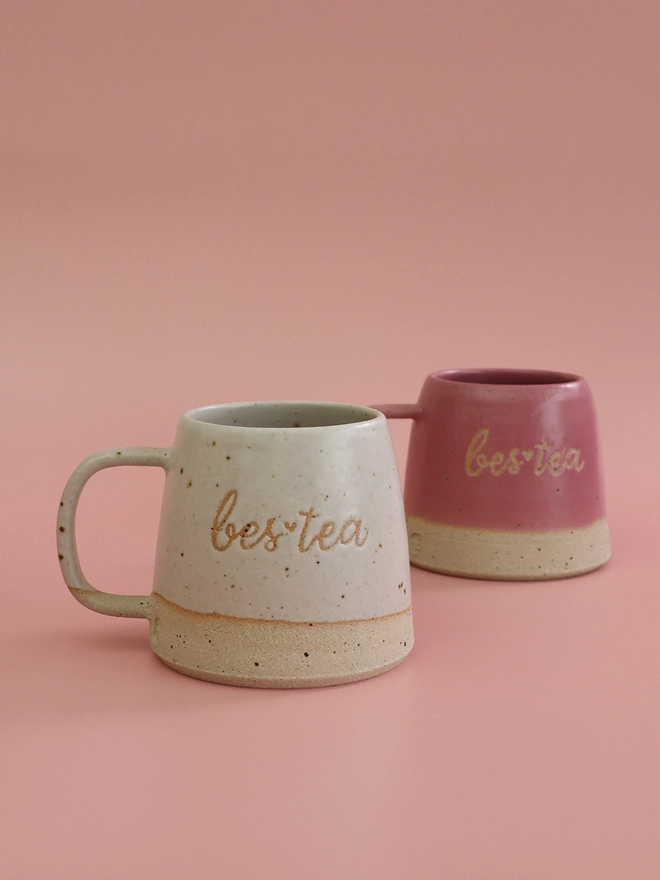 two handmade ceramic mugs with the saying bes-tea on each one infront of a pink background