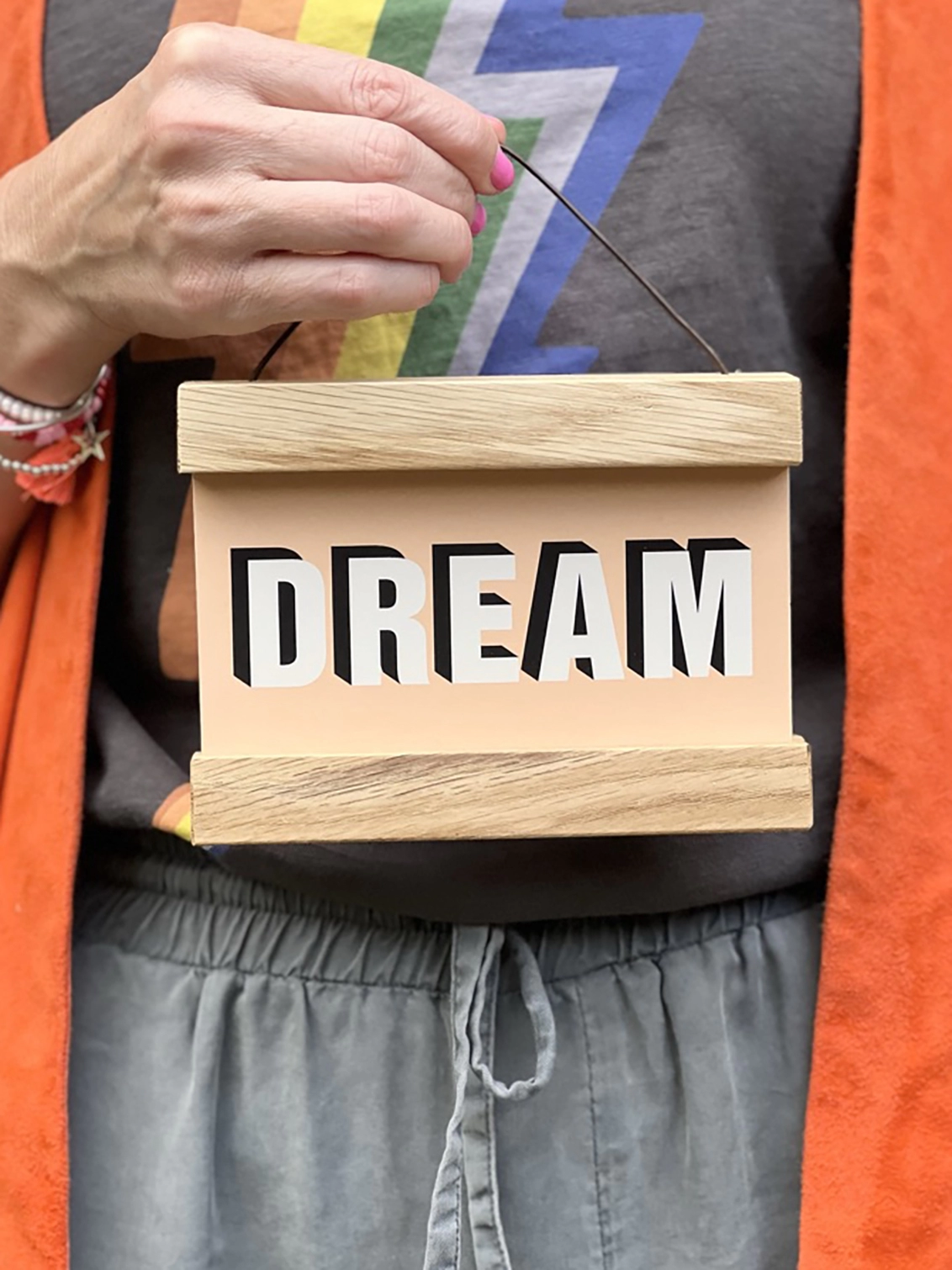 A print in peach colour with the word, DREAM framed in a oak hanger, with a waxed cotton cord for hanging being held by a hand with pink nail varnish 