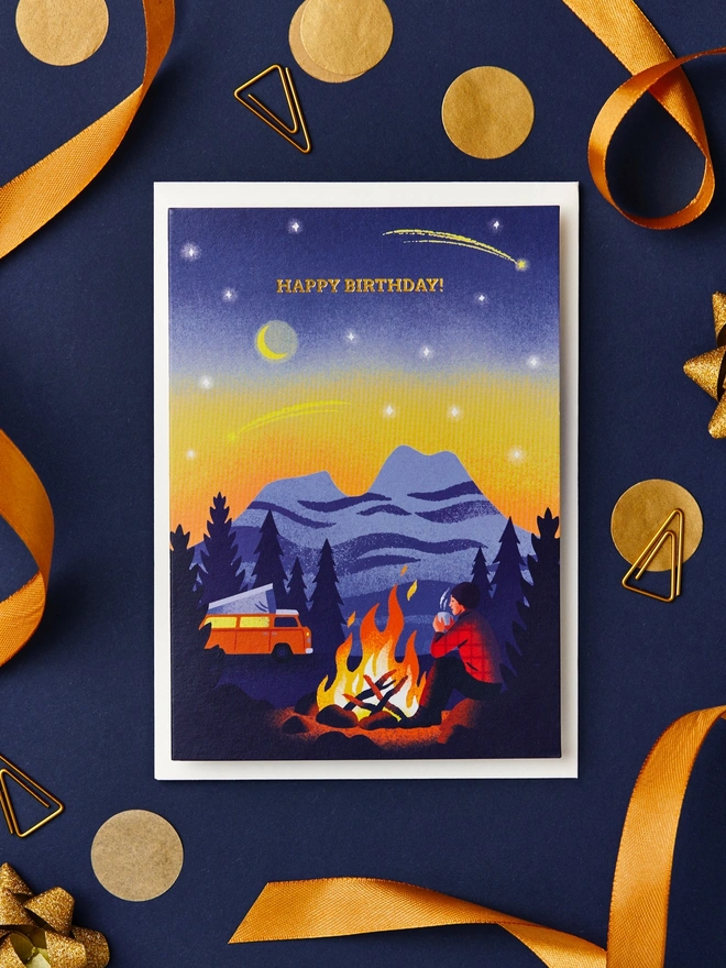 Greeting card with illustration of a camper sat beside a bonfire under a starlit sky. The card is on a desk with a navy blue background and gold ribbon decorations.