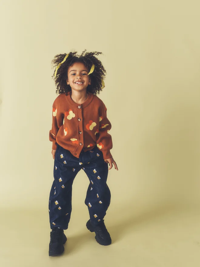 Child smiles mid-jump against a pale yellow backdrop, wearing a Studio Koter chestnut floral intarsia cardigan and dark denim trousers with Denim Petal embroidery.