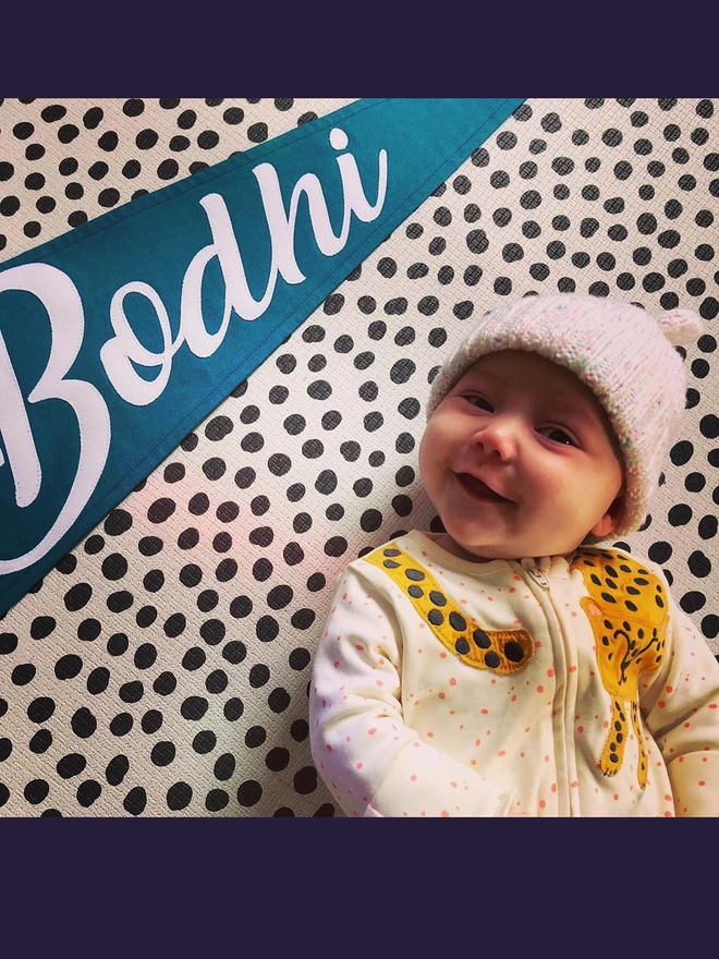 A pennant flag in teal with the name Bodhi in ivory laid next to a baby.