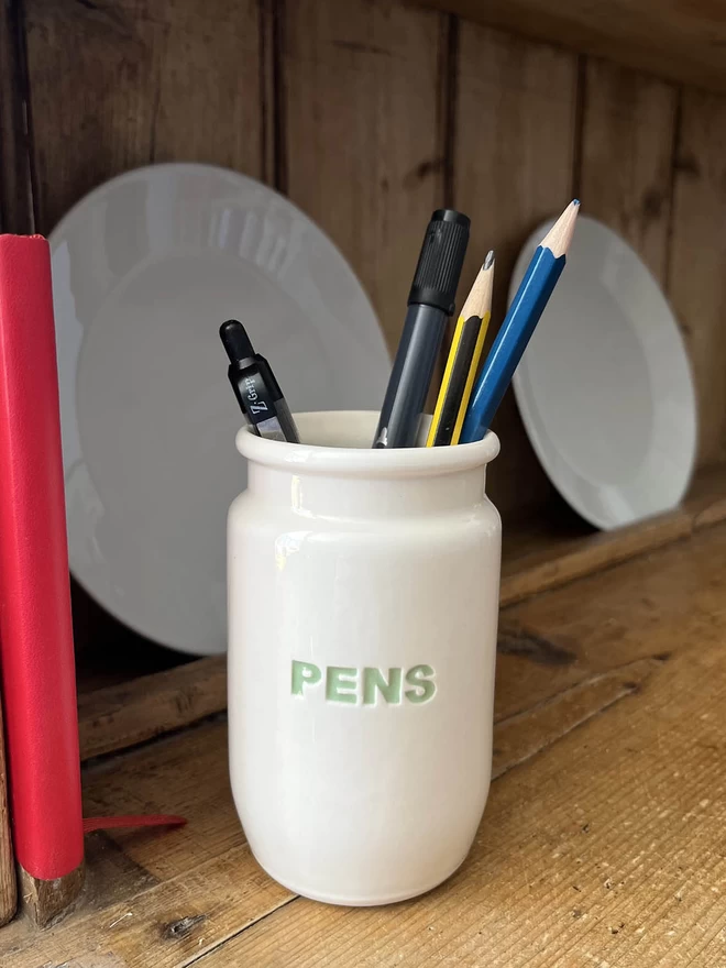 A handmade pot / jar has the word ‘pens’ in recessed green letters, holds an array of stationary. 