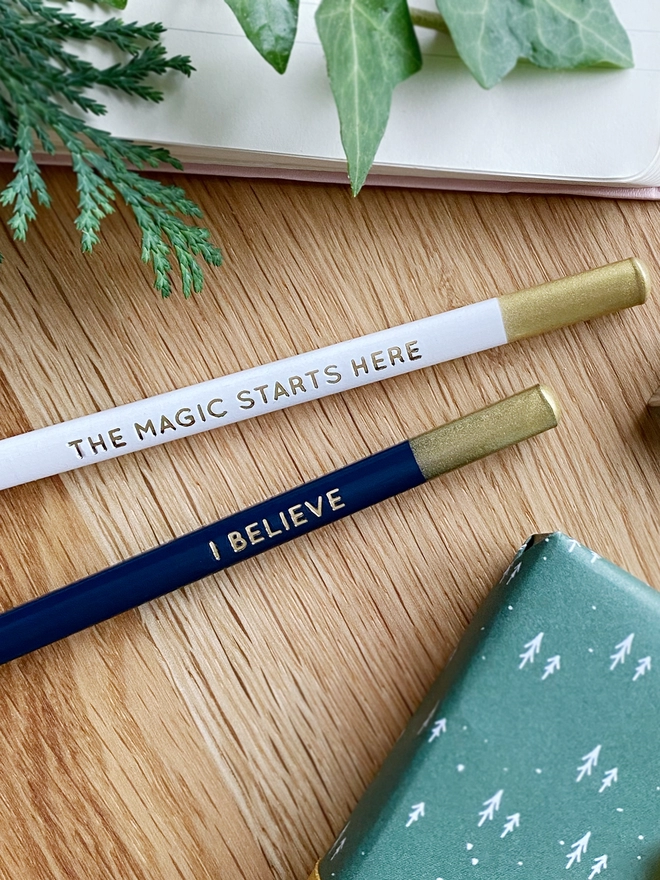 Two pencils, one white and one navy blue, both with gold writing along the sides, lay on a wooden desk.