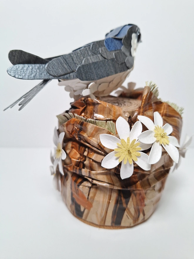 Housemartin bird sculpture, perched on a floral tree stump. This handmade sculpture is made from recycled paper. The bird is black and blue, with a white belly. White and yellow flowers adorn the tree stump. Back view.