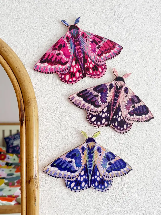 wooden moth wall decorations. pink, purple and blue moth decorations hung on plain white wall next to mirror