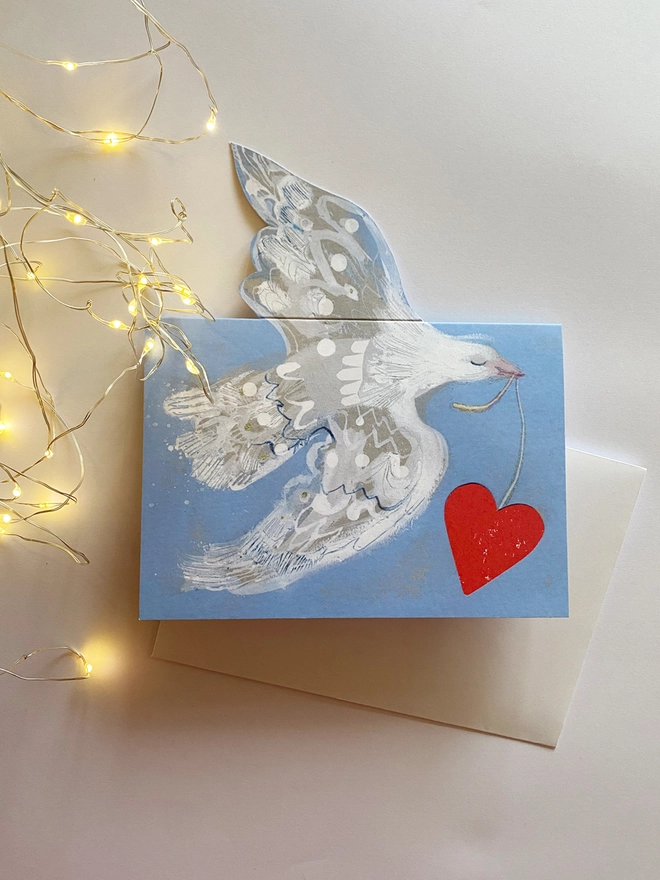 Esther Kent greetings card showing white flying dove in a light blue background, carrying a vibrant red heart on a string