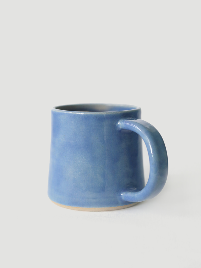 A handmade ceramic standard mug in glossy pacific blue glaze with a chunky loop handle, photographed against a plain light grey background