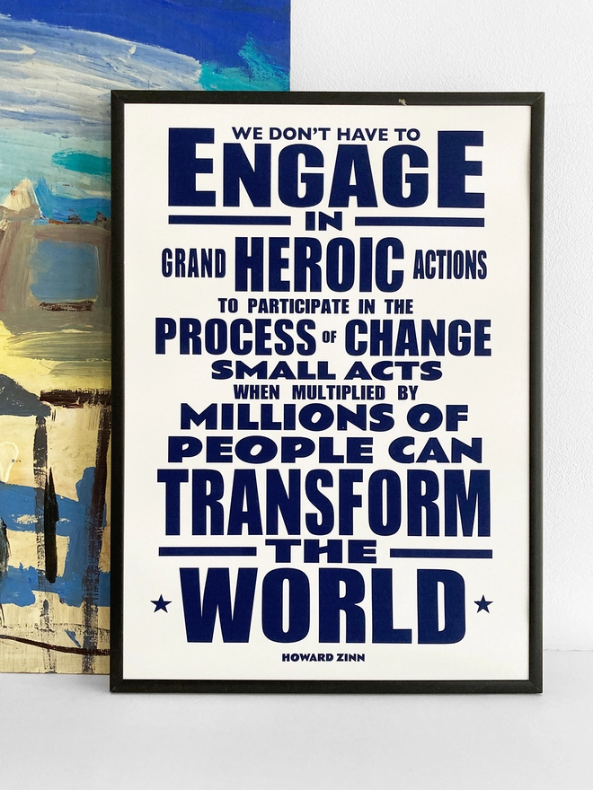 Framed typographic print, with navy blue text on a white background. Quote by Howard Zinn - “We don't have to engage in grand, heroic actions to participate in change. Small acts, when multiplied by millions of people, can transform the world.”    The print rests against a blue and yellow abstract painting.