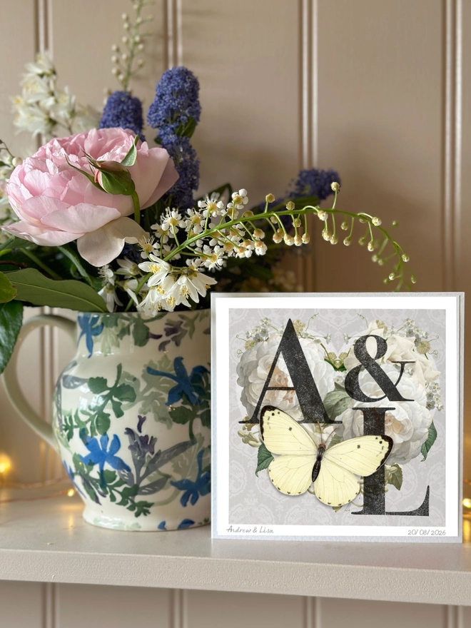 A handmade Diamond Wedding anniversary card featuring two initials of a couples names, place inside a heart design with vintage roses and on a silver grey background. The recipients initials are hand written on the front of card and hand signed, A white 3D papercut butterfly sits on the heart and wings can be folded out for display. The card is diplayed on a kitchen dresser next to a vase of flowers.