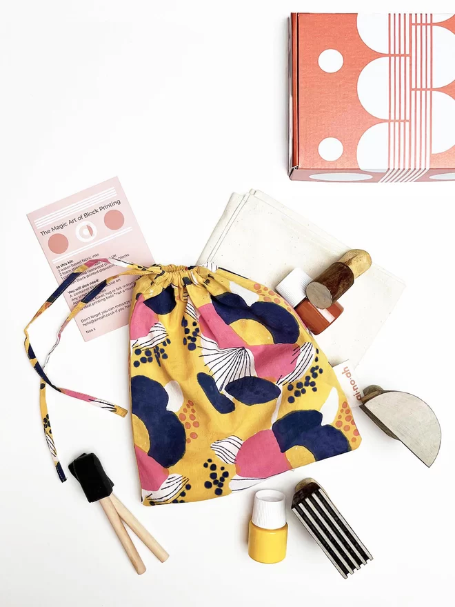 Block printing kit with a printed yellow drawstring bag, two paint pots, one yellow and one cinnamon coloured, two foam brushes, an instruction booklet, three geometric wooden blocks and a plain cream tea towel, next to a rectangular orange box with a white geometric design printed on it