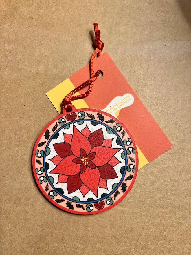 Multi coloured wooden tree decoration with a red poinsettia pattern at the centre. It has a red ribbon and is lying on a brown card background with a business card with Rebecca Strickson on it. 