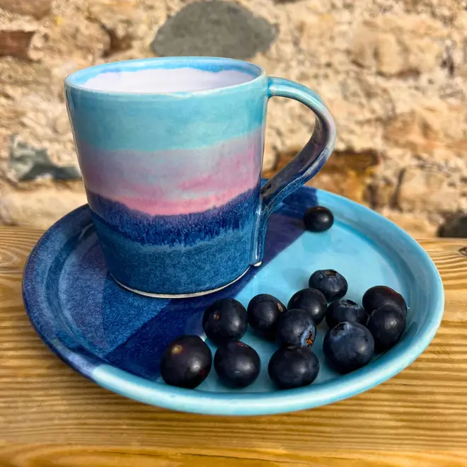 Landscape Standard Mug, a colourful mug placed on a wooden surface against a brick wall. It is accompanied by a colourful plate and some fruit. 
