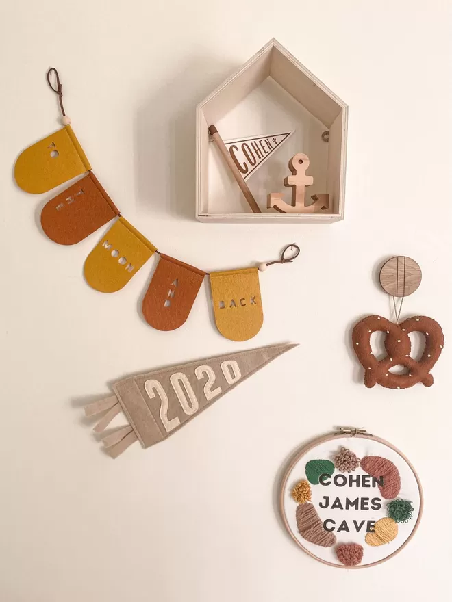 Garland in orange and mustard hanging on a cream wall with a 2020 pennant, a name plaque, a felt pretzel and wooden house shelf.