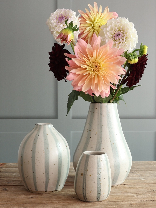 Handmade Ceramic Stoneware Vases in Blue and White Stripe in three different sizes - small, medium and large – sat on a wooden surface. The largest vase holds a bouquet of pink, yellow, white and red flowers.

