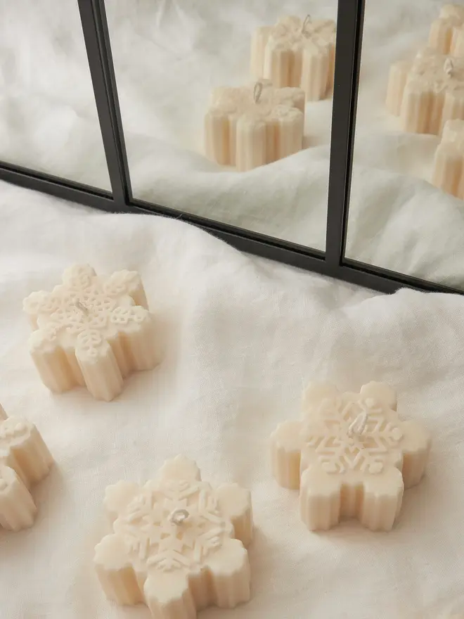 Classic Cream Snowflake Sculpture Candle. Cream snowflake-shaped candles with intricate embossed patterns and ribbed sides, arranged on white linen in front of a black-framed mirror. The delicate sculptural candles bring a soft, wintry touch to Christmas décor.