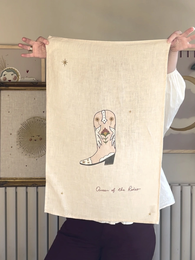 'Boots Are Made For Walking' Linen Tea Towel, a person holding up a linen tea towel with a boot motif on it. 