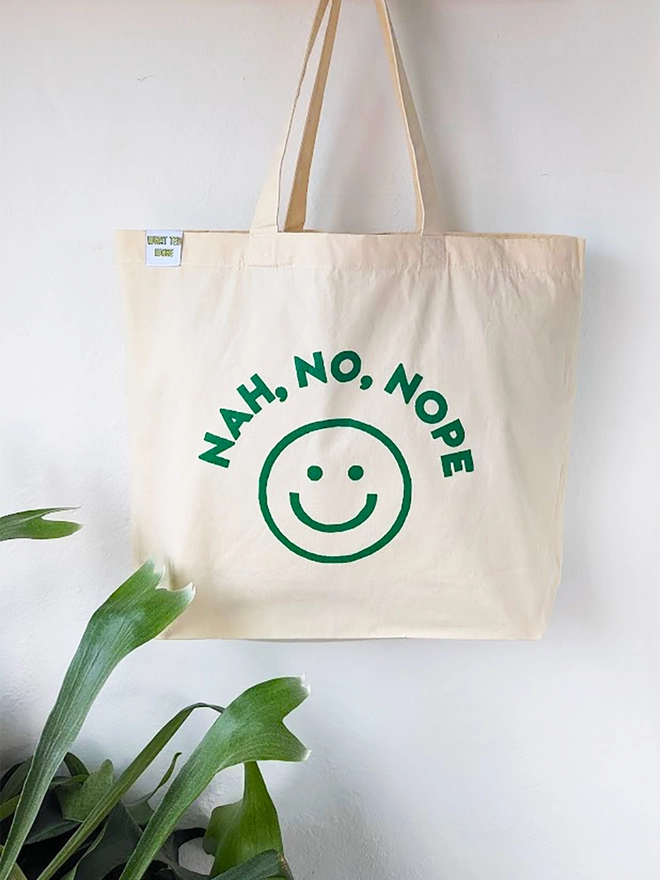 A hanging natural canvas tote bag with green Nah, No, Nope slogan and a graphic smiley face.