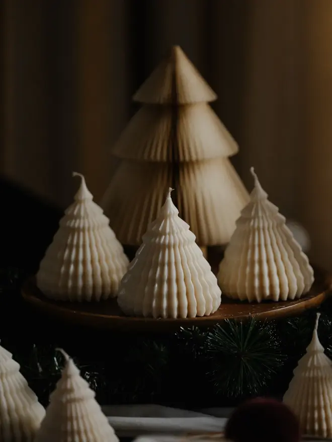 Origami Christmas Tree Candles (Set Of 2). A pair of glowing cream tree candles in two sizes against golden bokeh lights, gold and red baubles and pine greenery. The detailed ribbed finish makes them an elegant festive accent for a Christmas table.