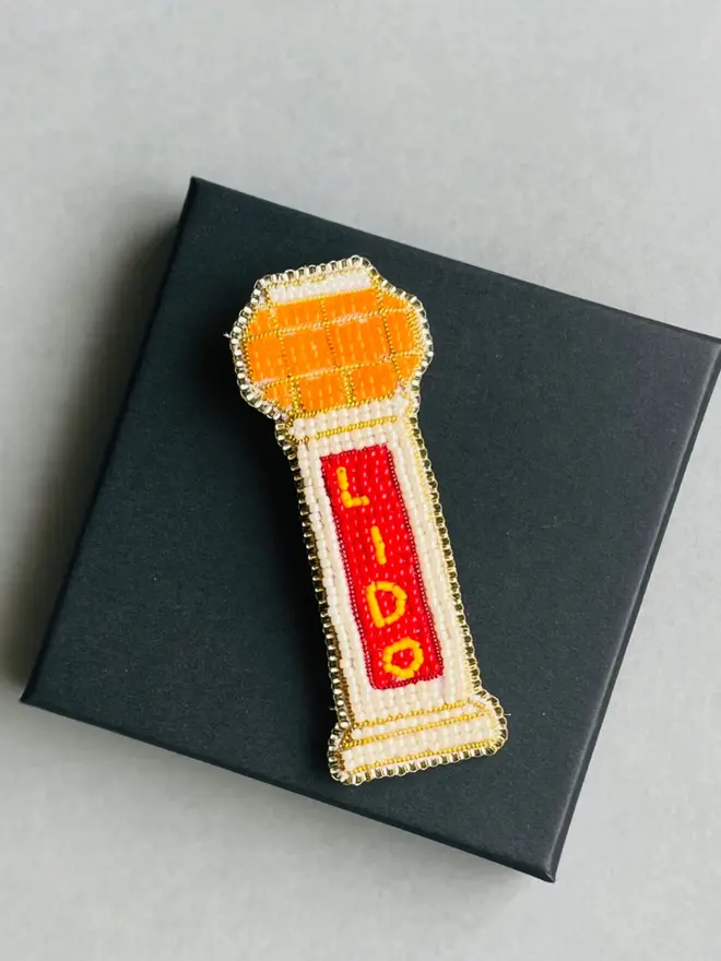 Beaded lido brooch with orange, red, and beige beads placed on top of a black box on a grey table.