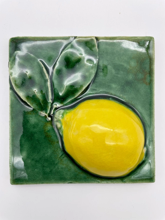 Handmade ceramic tile taken from a plaster cast of a real lemon, side view with two attached leaves. Very realistic, three-dimensional, with lush coloured glazes.