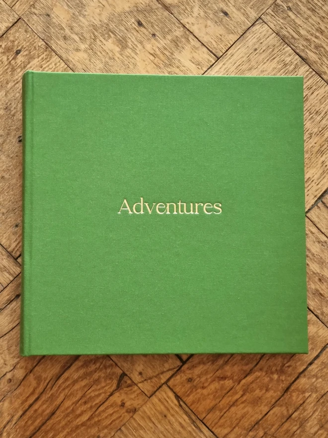 A hand-bound green photo album printed with the words 'Adventures' in gold lettering rests on a wooden floor.