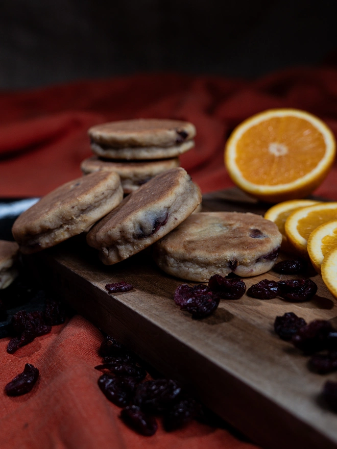 Orange and Cranberry Welsh Cakes on a wooden chopping board with loose cranberries an sliced orange
