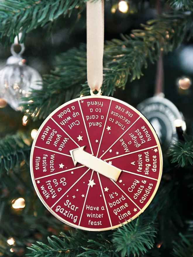 A deep red and gold Christmas decoration hangs on a Christmas tree by a gold ribbon. It has 12 segments, each one with a different Christmas activity idea, and a golden arrow in the centre.