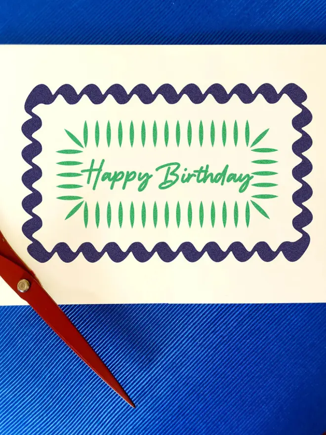 A yellow hand-printed birthday card with a blue wavy border and green decorative lines around the handwritten words “Happy Birthday,” displayed on a table.