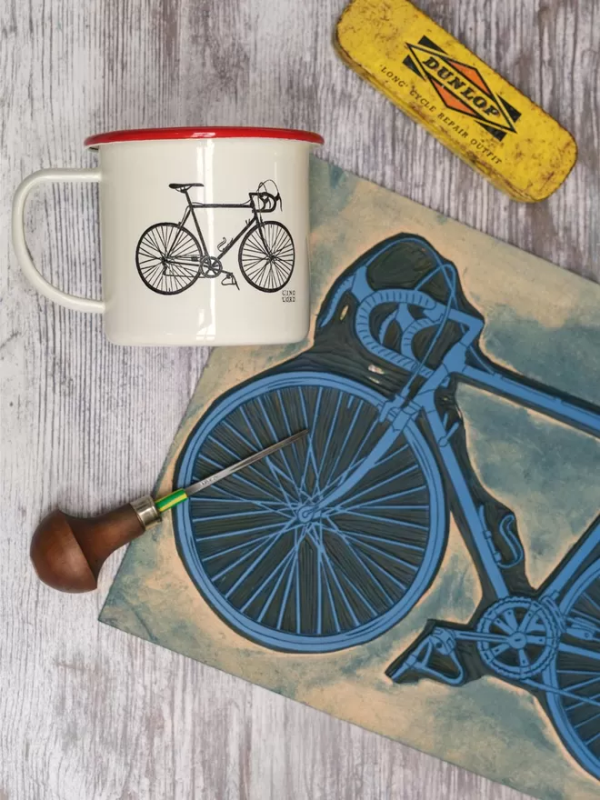 Picture of a Cream Enamel Mug with a Red Rim with a Bicycle design etched onto it, taken from an original Lino Print