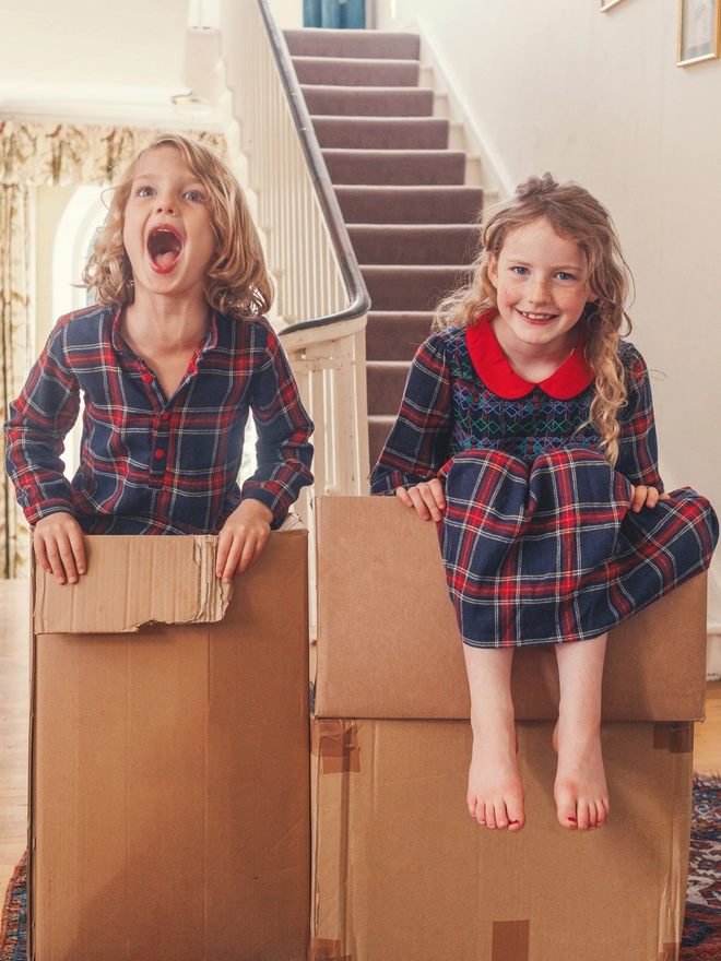 A boy and girl in matching navy tartan outfits hide in boxes