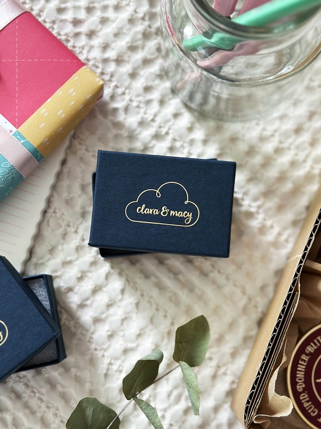 A navy blue gift box with the Clara and Macy logo embossed in gold on the lid rests on an ivory fabric surface.