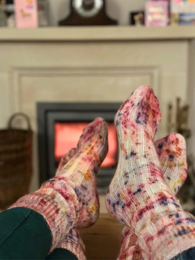 Two pairs of feet wearing studioEVIG's naturally dyed alpaca lounge socks, comfortably propped up on a table in front of a glowing fireplace, evoking warmth and relaxation