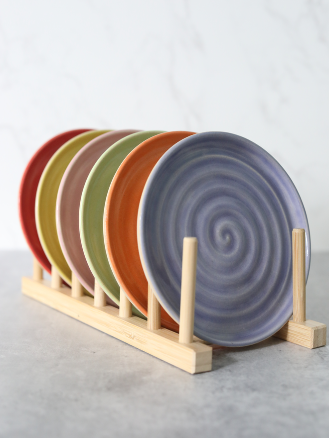 Five handmade ceramic cake plates displayed upright in a natural wood plate rack, fanned out to reveal a colour palette of cherry red, yellow, green, mandarin orange, and amethyst purple, each showing a concentric swirl texture, against a white marble background