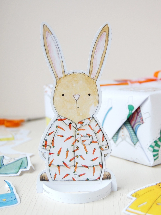 A gift wrapped in white wrapping paper with a hand illustrated outfit design and rabbit paper doll gift tags is beside a cut out rabbit paper doll on a white wooden table.