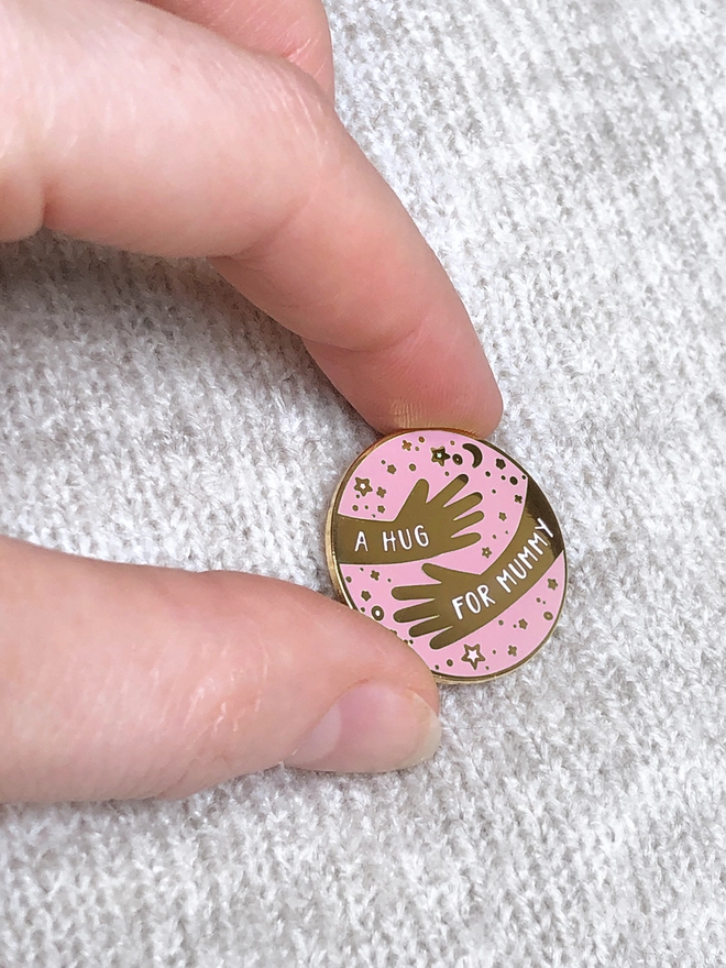 A hand is holding an enamel pin badge that has a hugging arms design and the words "A hug for Mummy".
