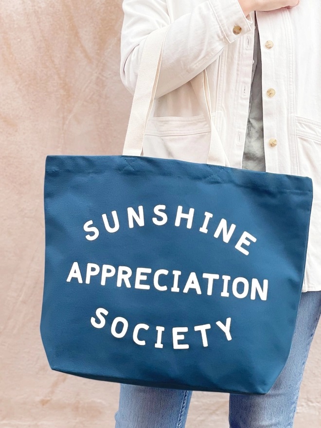 model carrying a large blue canvas tote bag with the words sunshine appreciation society on