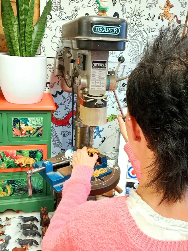 A hand holds a small Stegosaurus dinosaur on a clamp on the table of a pillar drill. A white tray of plastic animals sits below the pillar drill to the left. There is a small chest of drawers decorated in a brightly coloured jungle theme with animal drawer knobs attached with a snake plant in a white vase sitting on top. The dinosaur is being drilled and made into a dinosaur light switch by Candy Queen Designs.