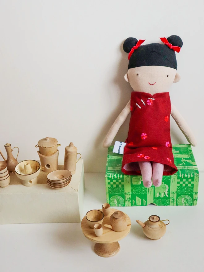 A handmade doll with black hair in buns, red bows, and a red dress with floral accents. A wooden horse and a book with an illustration are next to the doll. The background is a solid red.