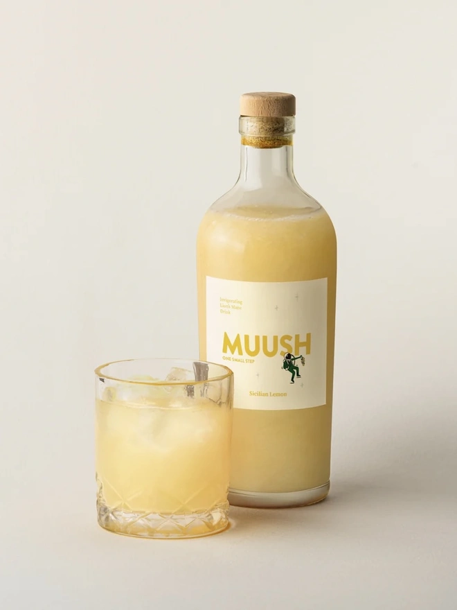 Sicilian Lemon Elixir, a bottle of soft drink sitting next to a glass of soft drink on a plain surface. 