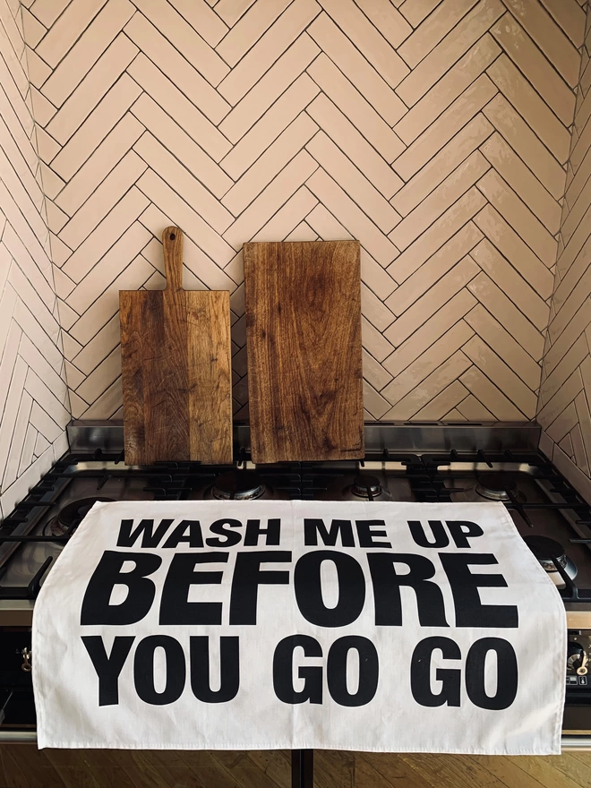 London Drying Wash Me Up Before You Go Go black screen printed text on white tea towel laying on oven with chopping boards in background