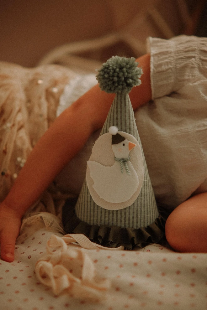 Baby Birthday Hat With Goose, a child lying with their birthday hat with a goose on a bed. 