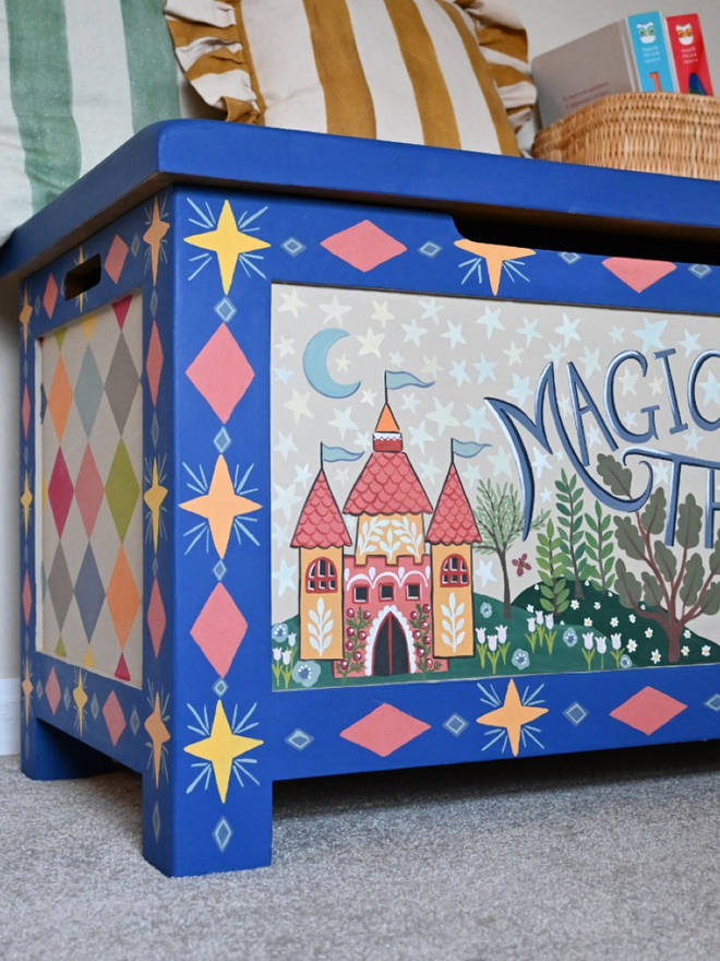 Close-up of a bright blue wooden chest with a hinged lid, painted with stars, diamonds, and whimsical illustrations including a pink castle with blue flags, mushrooms, and the words "Magical Things" in the center. Part of a striped pillow and a woven basket are visible in the top corner.