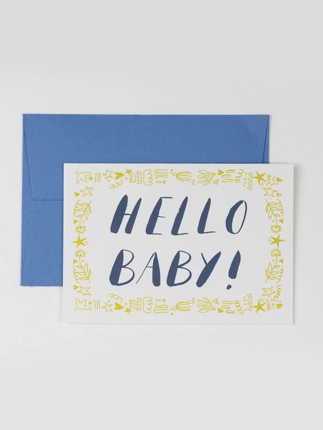A greetings card featuring a card that reads "Hello Baby!" in navy blue ink. The messaged is bordered with yellow pen and ink fine line pattern.