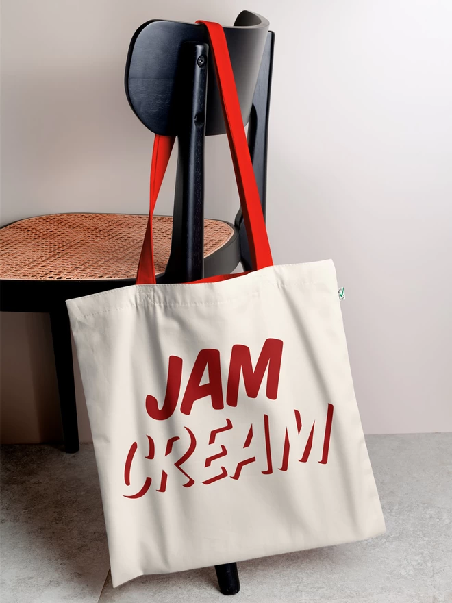 A white, natural coloured tote bag hanging on a chair, with the word with JAM written above CREAM in red ink - the bag is cream and the handles are red.