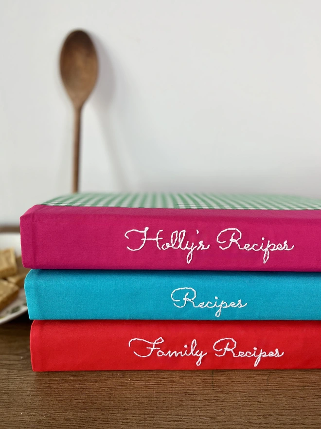 3 recipe binders with personalisation embroidered in white on pink, teal & red spines of the fabric covers, stacked on top of each other on wooden bench. Plate of baked ginger crunch squares and wooden spoon leaning against wall in background.
